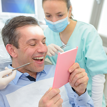 Older man checking smile in mirror while having lips pulled up by dentist