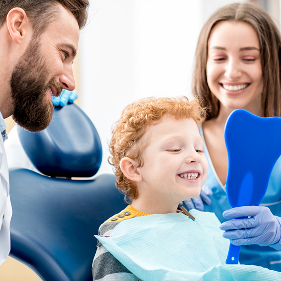 Adults next to child in dental chair showing him a mirror